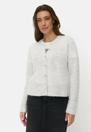 Kvinde med langt brunt hår iført en hvid strikket cardigan med knapper og et sort skørt, stående mod en ensfarvet lys baggrund.