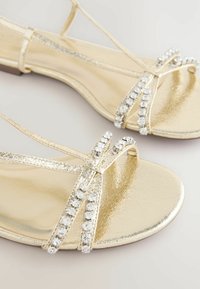 Sandalette dorate con finitura metallica lucida, caratterizzate da due cinturini curvi decorati con strass trasparenti sul davanti.