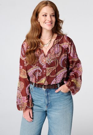 Vrouw met lang golvend rood haar, een kastanjebruine paisleyblouse en blauwe spijkerbroek, glimlachend en poserend met één hand in de zak.