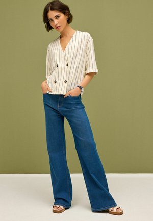 Gestreifte beige und weiße Bluse mit kurzen Ärmeln und doppelter Knopfschließe, kombiniert mit blauen Schlagjeans und hellbraunen Plateausandalen.