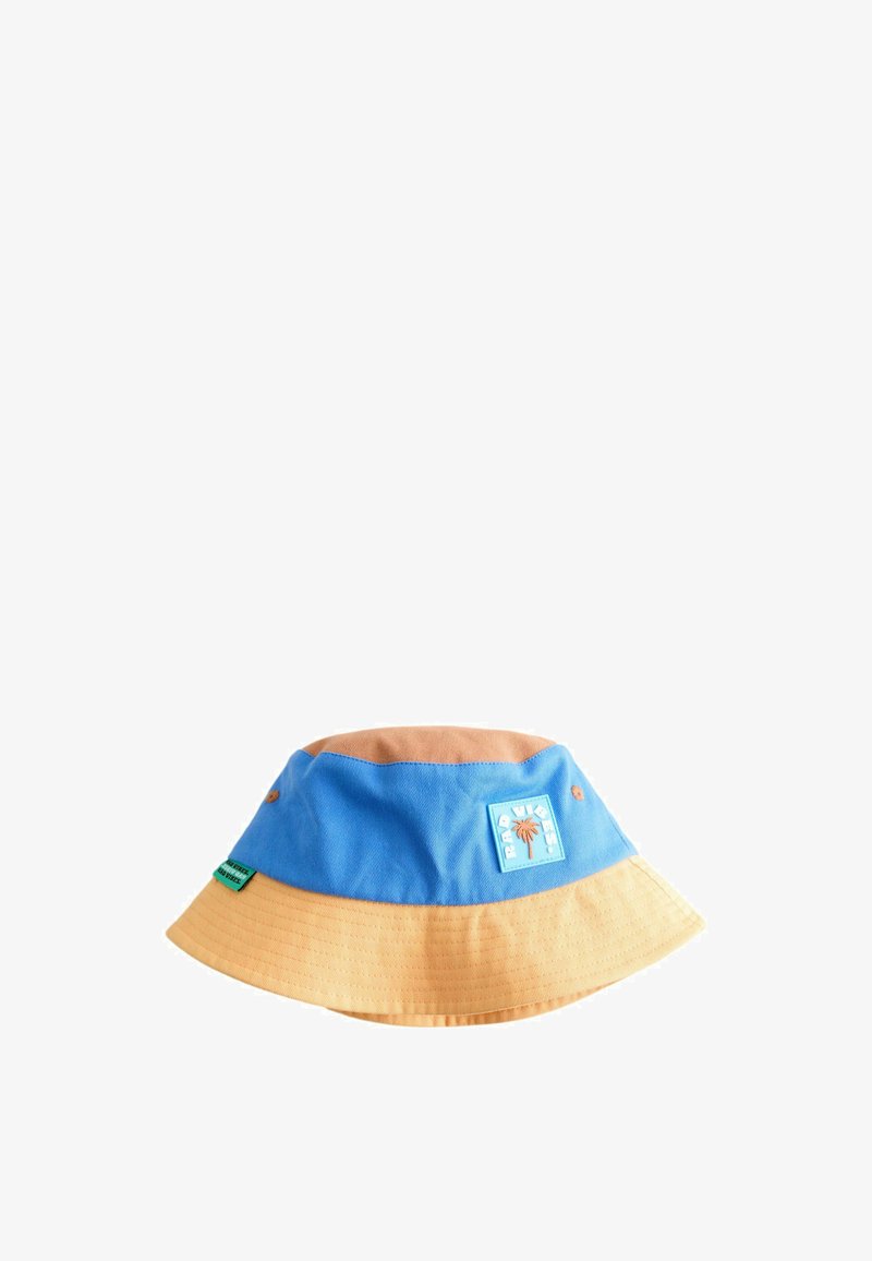Chapeau bob bicolore bleu et beige avec écusson brodé de palmier et petite étiquette verte sur le côté, sur fond blanc.