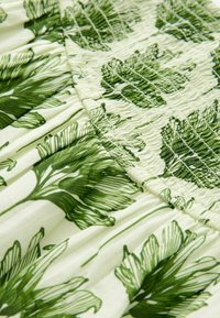 Groene stof met bladerenprint op een lichte achtergrond. Heeft gerimpelde details en een vloeiende textuur. Patronen van verschillende blaadvormen doorheen.