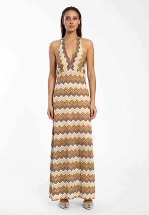 Frau trägt ein ärmelloses, braun-beiges Maxi-Kleid mit Zickzackmuster und Neckholder, kombiniert mit silbernen offenen High Heels, stehend vor weißem Hintergrund.