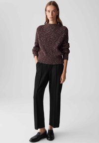 Frau in einem kastanienbraunen, gemusterten Strickpullover, schwarzer maßgeschneiderter Hose und schwarzen Slippern, die mit den Händen in den Taschen vor einem schlichten Hintergrund steht.