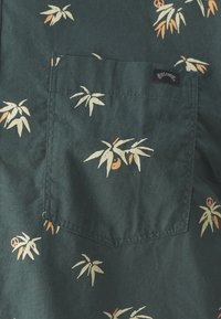 Chemise à manches courtes en tissu vert foncé, avec une poche et un motif répétitif de feuilles claires et de petites touches orange.