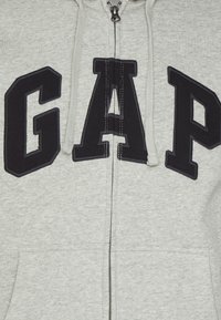 Grå zip-up hættejakke med store, mørkeblå "GAP" bogstaver over brystet, med en glat tekstur og hætte med snørre.