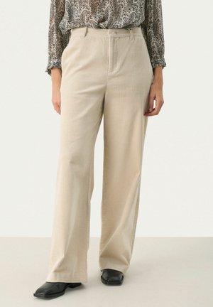 Beige Cord-Hose mit weitem Bein, hoher Taille und Gürtelösen, kombiniert mit schwarzen Schuhen mit quadratischer Spitze und einer gemusterten Langarmbluse.