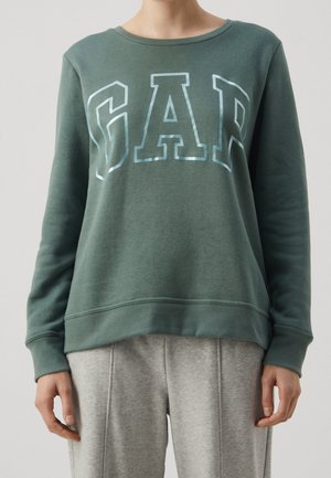 Person iført en grøn sweatshirt med reflekterende "GAP"-bogstaver og lysegrå sweatpants, stående mod en ensfarvet baggrund.