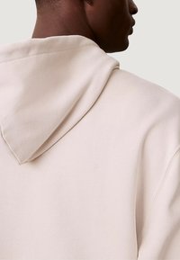 Felpa con cappuccio beige chiaro realizzata in tessuto morbido e strutturato, con cappuccio prominente e vestibilità comoda, dettagli di cuciture visibili e design pulito.