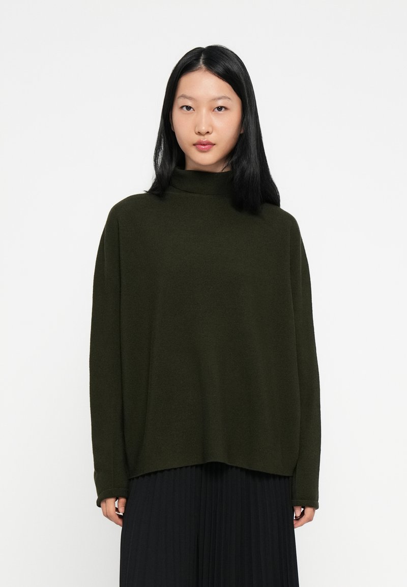 Donna con capelli neri lunghi che indossa un maglione a collo alto oversize verde scuro e una gonna plissettata nera, su uno sfondo bianco.