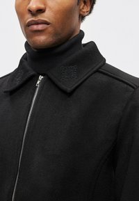 Crna jakna od mješavine vune s patentnim zatvaračem, špicastim ovratnikom i izvezenim cvjetnim ukrasima na ovratniku. Ima meku teksturu i strukturirani dizajn.