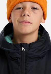 Jongere persoon die een oranje gebreide muts en een zwarte ritsjack met tealvoering draagt, met een neutrale uitdrukking en kijkend iets omhoog.