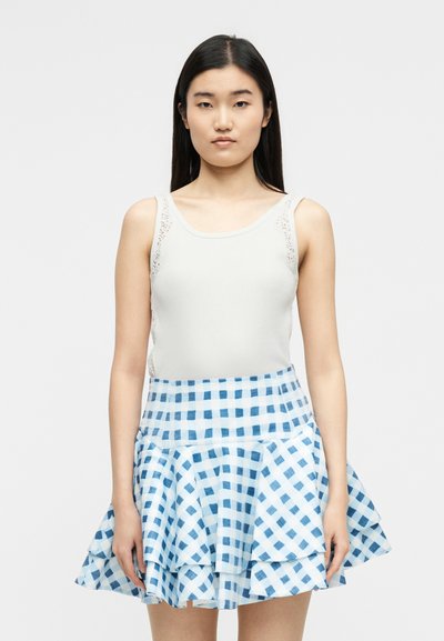 Γυναίκα με μακριά μαύρα μαλλιά που φοράει λευκό αμάνικο τοπ και μπλε-λευκή καρώ φούστα με στρώσεις, στέκεται μπροστά σε μια απλή φόντο.