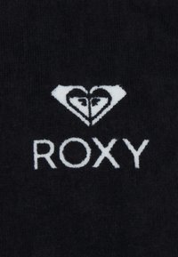 Musta kangas, jossa on valkoisella ommeltu logo, jossa on sydänkuvio ja tekstinä paksuilla, isoilla kirjaimilla "ROXY".