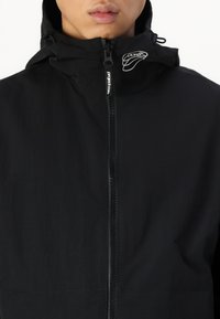 Czarna kurtka typu windbreaker z zamkiem błyskawicznym z przodu, z wysokim kołnierzem i akcentem logo. Materiał jest lekki o gładkiej strukturze.