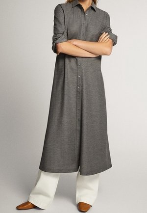 Personne portant une longue robe-chemise grise boutonnée avec manches retroussées, pantalon blanc à jambes larges et chaussures marron pointues, debout les bras croisés.