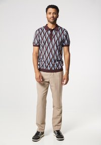 Polo de punto de manga corta en un patrón ondulado marrón, azul y blanco, con un cuello oscuro y cierre de botones. Combinado con pantalones claros color beige y zapatillas negras.