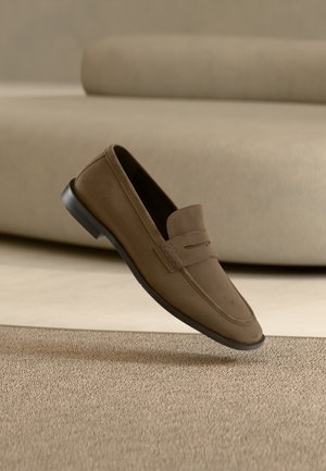Mocassin en daim marron incliné au-dessus d'un tapis beige avec un canapé beige assorti en arrière-plan flou.