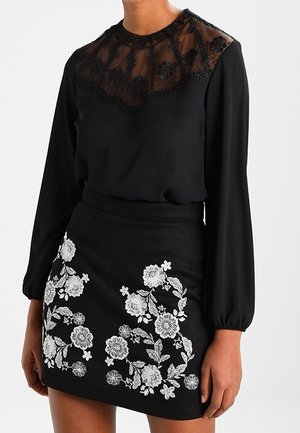 Femme portant un chemisier noir à manches longues avec un empiècement en dentelle transparente et une jupe noire brodée de motifs floraux blancs.