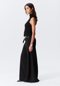 Haut noir court sans manches à col montant, associé à un pantalon noir à jambes larges avec une ceinture à œillets métalliques. Détail bracelet argenté.