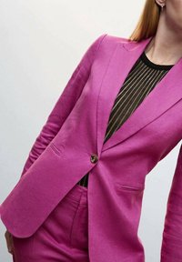 Blazer fuchsia sur mesure avec une fermeture à bouton unique, des revers à encoche et des poches passepoilées, porté sur un haut rayé noir. Texture de tissu lisse.