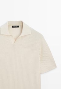 Polo en maille côtelée à manches courtes couleur crème avec un col doux et une petite étiquette de marque noire à l’intérieur de l’encolure.