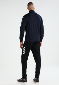 Marineblauwe sportjack met een staande kraag, gecombineerd met zwarte broeken met witte chevronaccenten. Model draagt zwarte voetbalschoenen.