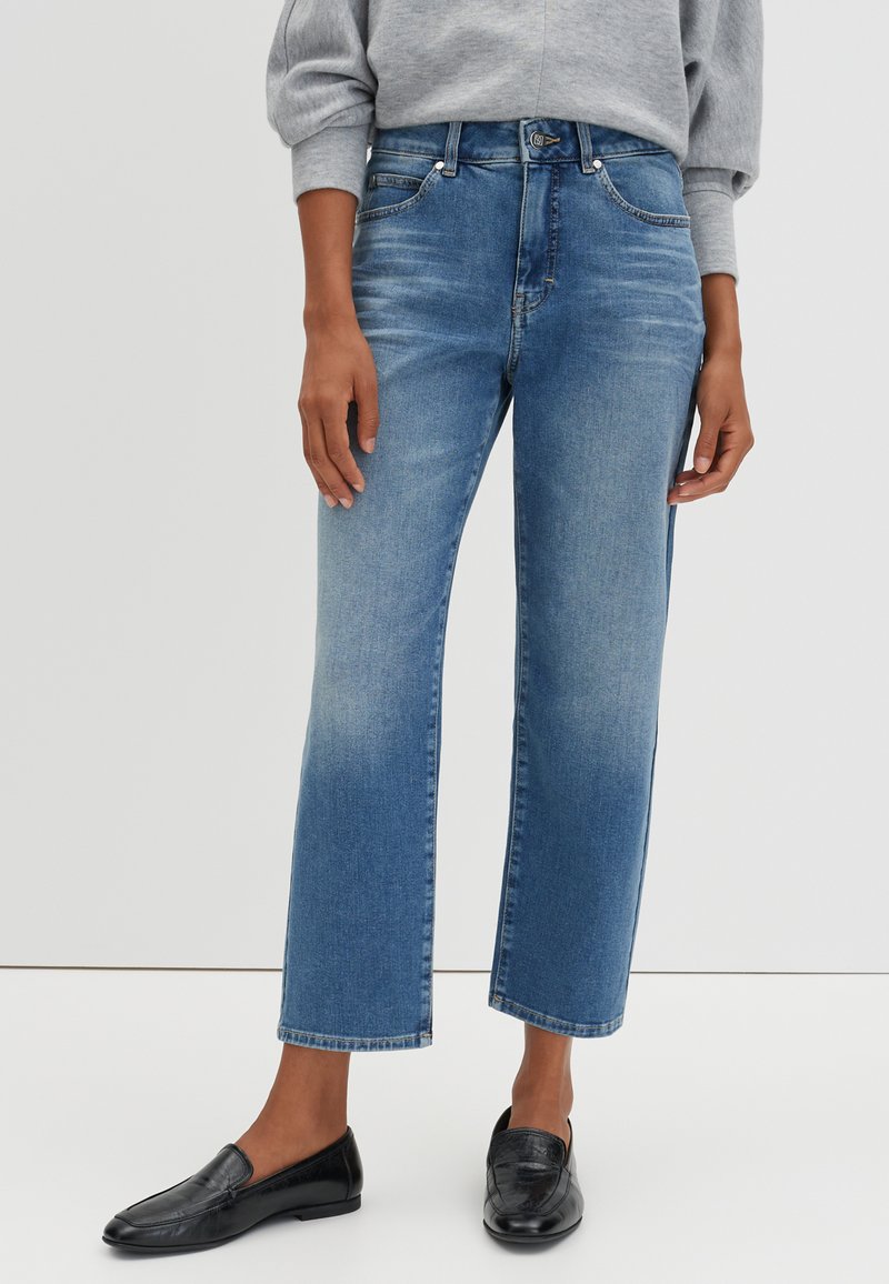 Jeans cropped bleus mi-hauteurs avec une légère décoloration, portés avec des mocassins noirs et un sweat-shirt gris sur une personne debout devant un fond uni.