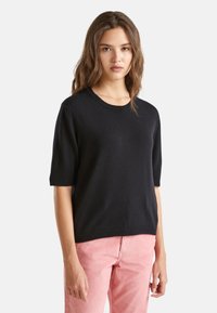 Młoda kobieta z falującymi brązowymi włosami, ubrana w czarny sweter z krótkim rękawem i różowe spodnie z kordurowej tkaniny, stojąca na jednolitym tle.