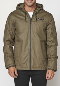 Mężczyzna w oliwkowej zielonej kurtce z kapturem, z zamkiem błyskawicznym z przodu i dwiema kieszeniami po bokach, zestawionej z czarnymi spodniami.