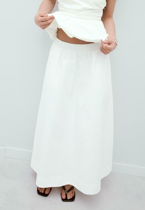 Žena oblečená v dlhej bielej sukni, mierne zdvíhajúca biely top, stojaca bosá na svetlej podlahe oproti hladkej stene.