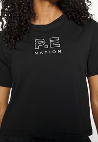 Black cotton t-shirt with a round neckline and short sleeves, featuring a white graphic print of "P.E NATION" on the front.