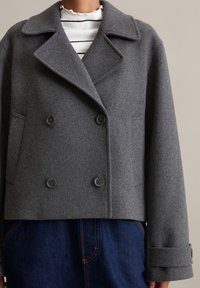 Dubbelbreasted grijze wollen jas met grote revers, twee voorkruike zakken en zwarte knopen. Draagt over een wit gestreepte top en donkere denim.
