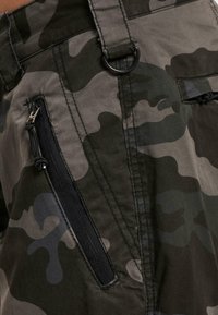Tissu camouflage avec une poche zippée noire verticale et une sangle cousue tenant un anneau en D en métal sur un vêtement en gros plan.