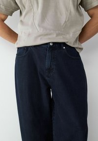 Ciemnoniebieskie dżinsy o luźnym kroju, pięciokieszeniowym designie i metalowym zapięciu. Materiał ma gładką teksturę i subtelny połysk.