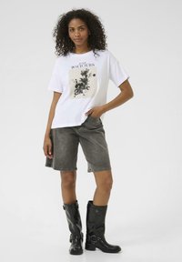 T-shirt blanc avec un motif floral, associé à un short en denim gris. Bottes noires montantes avec des sangles et des boucles, texture lisse.