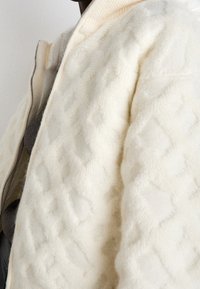 Vit ulljacka med en strukturerad diamantmönster, med dragkedja och ribbad krage. Mjuk finish och väldefinierad design.