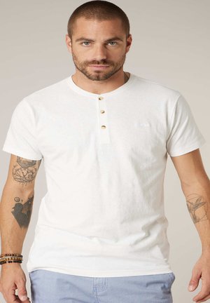 Man met kort haar en baard, gekleed in een wit henleyshirt met korte mouwen en een blauwe broek, met tatoeages op beide armen en armbanden aan de linkerpols.