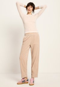 Eine Frau posiert mit erhobenen Armen, trägt ein transparentes Langarmshirt, beigefarbene weit geschnittene Hose und rosa Turnschuhe mit dunkelblauen Details.