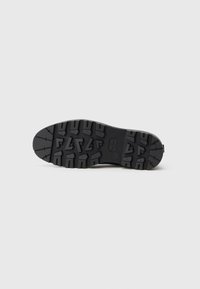 Suola di scarpa nera e robusta con scanalature profonde, modelli angolari rettangolari e triangolari, su uno sfondo chiaro e uniforme.