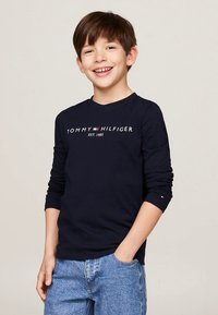 Mørkeblå langærmet skjorte med "TOMMY HILFIGER EST. 1985" trykt i hvidt og rødt, kombineret med lyseblå denimjeans. Glat tekstur.