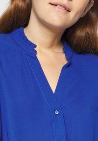 Blusa blu con colletto arricciato, scollo a V e dettagli con bottoni. Tessuto liscio con una leggera lucentezza e cuciture a contrasto.