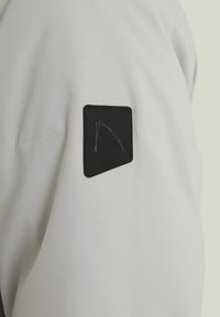 Ljust grått tyg med en slät textur har en framträdande svart rektangulär patch med en logotyp, placerad på axelområdet av plagget.