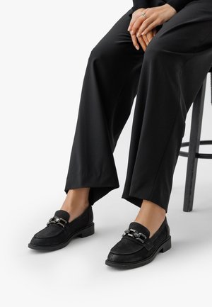 Mocasines de ante negro con un bajo tacón cuadrado, que presentan un acento decorativo de cadena de metal y una puntera redondeada, mostrados con unos pantalones anchos negros.