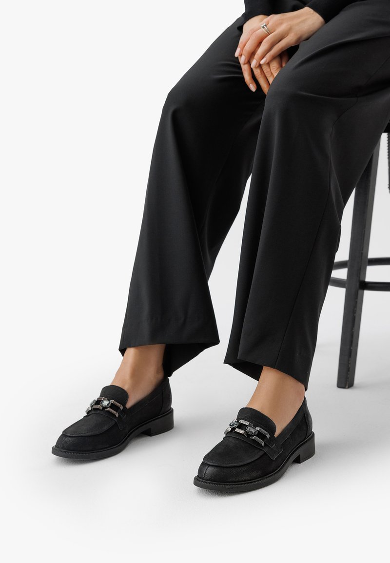 Mustadest sametist loafers madalate plokk-kontidega, kus on dekoratiivne metallketi aksessuaar ja ümar nina, esitatud mustade laiade pükstega.