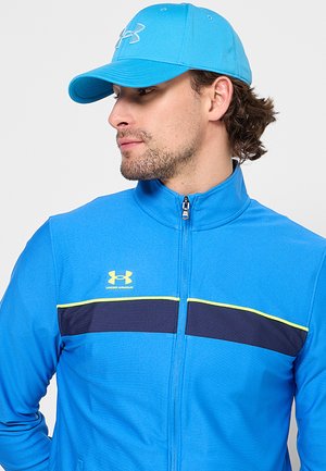 Man wearing blue Under Armour zip-up jacket and matching blue cap looking sideways against a plain background.