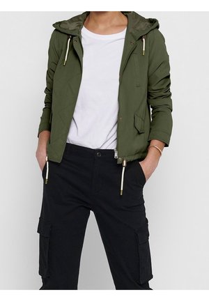 Personne portant une veste à capuche vert olive non fermée sur un t-shirt blanc et un pantalon cargo noir, les mains dans les poches.