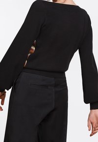 Pull en maille noir à manches longues, avec un motif coloré sur les côtés. Associé à un pantalon noir ajusté, présentant une texture lisse.