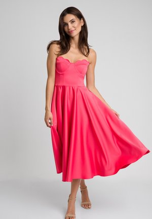 Fuksjowa sukienka bez ramiączek z falbaniastym korpusem, zwiewną spódnicą w kształcie litery A i gładką teksturą, zestawiona z cielistymi szpilkami.