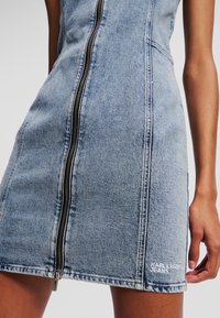 Jeans obleka s sprednjim zadrgo v svetlo modri barvi, ki ima izšito logotip v spodnjem levem kotu in vidne šive ob šivih.